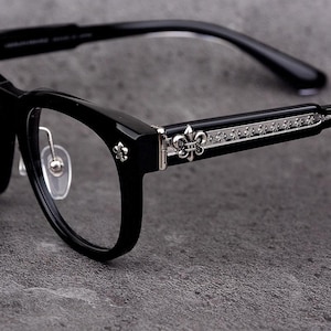 以下が含まれることがあります： 長方形のフレームとシルバーの装飾が施された黒い眼鏡。テンプルには、小さな繰り返し模様とヒンジ近くのフルール・ド・リスのエンブレムが付いた装飾的なシルバーバンドが付いています。レンズはクリアです。