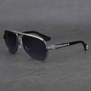 Puede incluir: Gafas de sol estilo aviador con montura plateada y lentes oscuras. Las patillas negras presentan detalles decorativos plateados, incluyendo flor de lis y detalles ornamentados. Las gafas tienen doble puente y un dise&ntilde;o elegante.