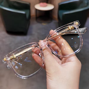 Op de afbeelding: Transparante bril met een vierkant montuur en gouden accenten. De veren en de brug hebben gouden details. De glazen zijn helder. De bril wordt door een hand vastgehouden. Het ontwerp is modern en elegant.