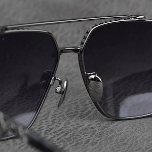 Puede incluir: Primer plano de unas gafas de sol con montura de metal plateado y lentes oscuras. Las gafas de sol tienen un doble puente y detalles decorativos en el borde superior. Las almohadillas nasales son plateadas y presentan un emblema decorativo.
