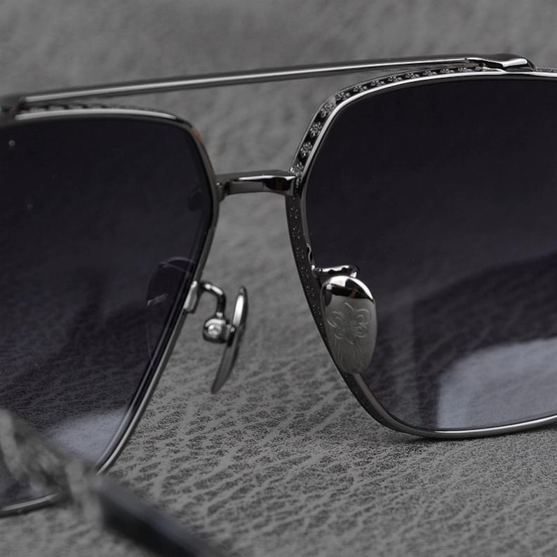 Puede incluir: Primer plano de unas gafas de sol con montura de metal plateado y lentes oscuras. Las gafas de sol tienen un doble puente y detalles decorativos en el borde superior. Las almohadillas nasales son plateadas y presentan un emblema decorativo.