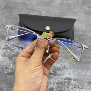 Op de afbeelding: Zilverkleurige bril met heldere glazen. De bril heeft een rechthoekige vorm en wordt voor een zwart leren etui gehouden. Het montuur is gemarkeerd met "B-TITANIUM".