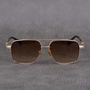 Puede incluir: Gafas de sol doradas con lentes degradadas marrones. Las gafas tienen un doble puente y lentes cuadradas. Las patillas son de color marr&oacute;n oscuro y presentan detalles decorativos. El dise&ntilde;o es moderno y elegante.