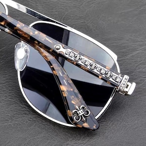 Puede incluir: Gafas de sol con montura plateada y lentes oscuras. Las patillas presentan un patr&oacute;n de carey marr&oacute;n y detalles plateados ornamentados. Una flor de lis plateada est&aacute; en la bisagra de la patilla.