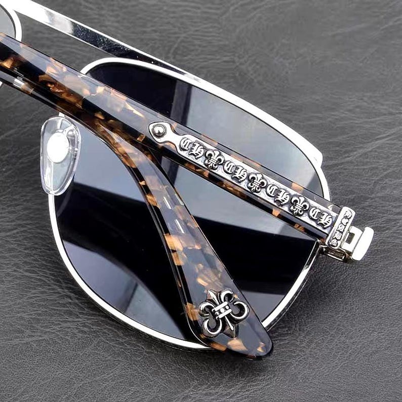 Puede incluir: Gafas de sol con montura plateada y lentes oscuras. Las patillas presentan un patr&oacute;n de carey marr&oacute;n y detalles plateados ornamentados. Una flor de lis plateada est&aacute; en la bisagra de la patilla.