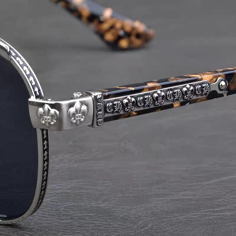 Puede incluir: Primer plano de gafas de sol con montura plateada y lentes oscuras. Las patillas presentan un patr&oacute;n de carey y detalles decorativos plateados, incluyendo emblemas de flor de lis. El dise&ntilde;o es intrincado y elegante.