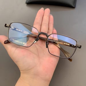 Op de afbeelding: Rechthoekige bril met een titanium montuur. De bril heeft een donkerbruin en zilver kleurenschema, met de tekst "TITANIUM IP" zichtbaar op de lens. De pootjes van de bril zijn lichtbruin.