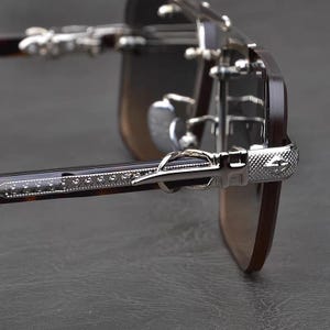 Puede incluir: Primer plano de gafas de sol con montura y lentes de color marr&oacute;n oscuro. Las patillas presentan un dise&ntilde;o de carey y est&aacute;n adornadas con detalles met&aacute;licos plateados. Las bisagras y las puntas de las patillas tambi&eacute;n son plateadas.