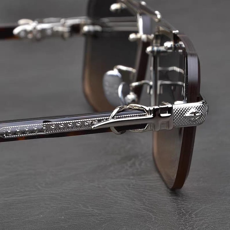Puede incluir: Primer plano de gafas de sol con montura y lentes de color marr&oacute;n oscuro. Las patillas presentan un dise&ntilde;o de carey y est&aacute;n adornadas con detalles met&aacute;licos plateados. Las bisagras y las puntas de las patillas tambi&eacute;n son plateadas.