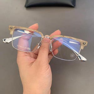 Op de afbeelding: Bril met een licht houten frame en zilveren accenten. De lenzen zijn rechthoekig met een zilveren bovenbalk. De tekst "TITANIUM IP" is zichtbaar op de lens. De veren hebben een decoratief ontwerp.