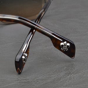 Puede incluir: Primer plano de gafas con un patr&oacute;n de carey marr&oacute;n. Las patillas presentan un emblema plateado de flor de lis. Las gafas tienen un dise&ntilde;o elegante y moderno, con un enfoque en los detalles de la montura.