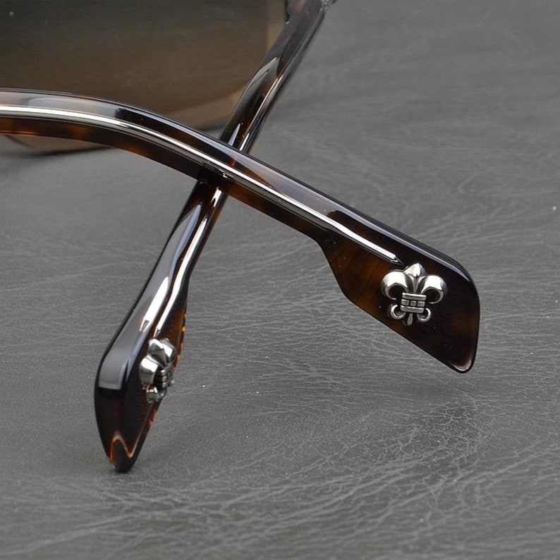 Puede incluir: Primer plano de gafas con un patr&oacute;n de carey marr&oacute;n. Las patillas presentan un emblema plateado de flor de lis. Las gafas tienen un dise&ntilde;o elegante y moderno, con un enfoque en los detalles de la montura.
