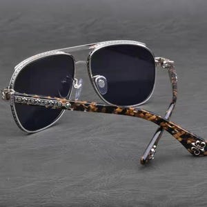 Puede incluir: Gafas de sol estilo aviador con lentes oscuras y montura plateada. Las patillas presentan un patr&oacute;n de carey marr&oacute;n y negro. La montura est&aacute; adornada con peque&ntilde;os detalles decorativos.