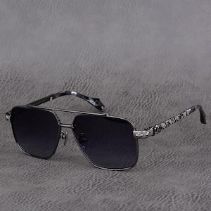 Puede incluir: Gafas de sol grises oscuras con doble puente y montura cuadrada. Las lentes tienen un degradado de oscuro a claro. Las patillas tienen un estampado de camuflaje y detalles met&aacute;licos decorativos. Las gafas est&aacute;n sobre un fondo gris.