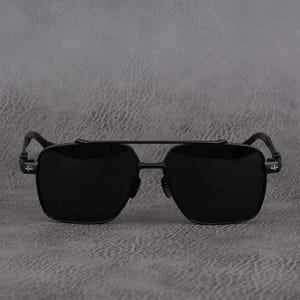 Puede incluir: Gafas de sol negras con doble puente y lentes cuadrados. Las monturas son de metal negro con un dise&ntilde;o de cruz en las patillas. Las gafas de sol est&aacute;n sobre un fondo gris.