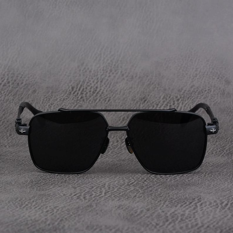 Puede incluir: Gafas de sol negras con doble puente y lentes cuadrados. Las monturas son de metal negro con un dise&ntilde;o de cruz en las patillas. Las gafas de sol est&aacute;n sobre un fondo gris.