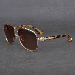 Puede incluir: Gafas de sol de aviador con montura dorada y lentes marrones. Las patillas presentan un estampado de carey y detalles dorados. Las gafas tienen doble puente y un dise&ntilde;o cl&aacute;sico.