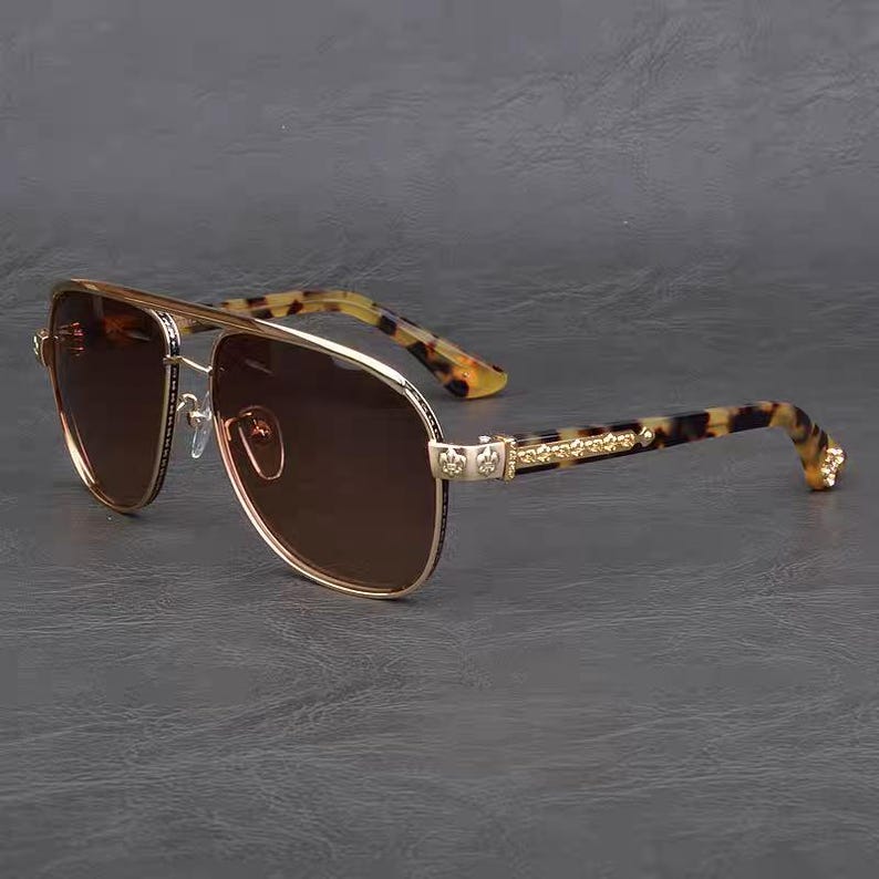 Puede incluir: Gafas de sol de aviador con montura dorada y lentes marrones. Las patillas presentan un estampado de carey y detalles dorados. Las gafas tienen doble puente y un dise&ntilde;o cl&aacute;sico.