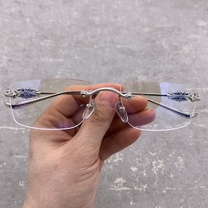 Op de afbeelding: Bril zonder montuur met zilverkleurige metalen montuur. De veren en de hoeken van de glazen hebben een decoratief bloemmotief. De glazen zijn helder en de bril wordt door een hand tegen een grijze achtergrond gehouden.