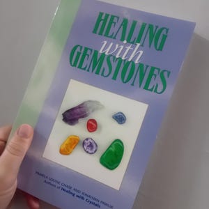 Puede incluir: Un libro titulado "Healing with Gemstones" con una portada de color púrpura claro. La portada presenta el título en verde y blanco, y una imagen de varias piedras preciosas. Los nombres de los autores están listados en la parte inferior.