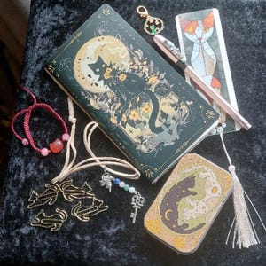 May include: A collection of cat-themed stationery and accessories. Features a journal with a black cat illustration, a pen, a bookmark, a small tin, a bracelet, and cat-shaped paper clips. The items are in shades of green, gold, and pink, arranged on a dark background.