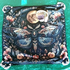 May include: A square, dark blue decorative cloth featuring a large moth illustration with intricate details. The moth is surrounded by flowers, a full moon, and a shell with amethyst crystals. The corners have white lace trim.