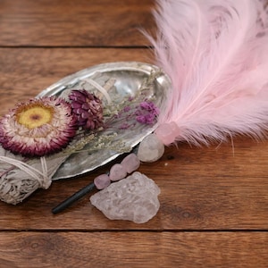 Peut inclure: Une composition d'objets spirituels sur une surface en bois. Comprend un paquet de sauge, des fleurs séchées, une plume, des cristaux et une coquille. Les fleurs sont violettes et jaunes, et la plume est rose. Les cristaux sont de formes et de tailles variées.