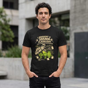 May include: A black t-shirt featuring a graphic of a detective using a 395NM flashlight to examine glassware. The text reads: I BROUGHT A 395NM FLASHLIGHT. THIS IS NOW AN INVESTIGATION. The illustration includes green glowing glass items and magnifying glasses.
