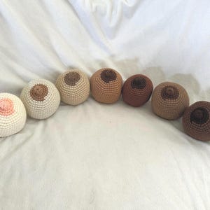 May include: A collection of seven crocheted breast forms in varying skin tones. The forms are arranged in a semi-circle, showcasing a range of colors from light pink to dark brown. Each form has a detailed nipple and areola.