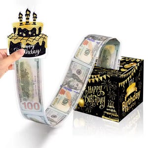 May include: A black and gold birthday gift box with a money pull-out feature. The box is decorated with fireworks and the words "Happy Birthday". A decorative cake topper with candles and the words "Happy Birthday" is attached to the money.