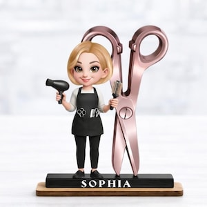Può includere: Una statuina di un parrucchiere che tiene un asciugacapelli nero e una spazzola, con grandi forbici color oro rosa dietro di lei. Indossa un grembiule nero con icone di forbici e pettine, pantaloni neri e scarpe nere. La base recita "SOPHIA".