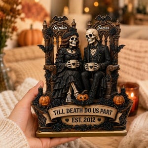 May include: A decorative figurine featuring two skeletons dressed in formal black attire, seated on ornate thrones. The piece includes the names Sophia and James, the text TILL DEATH DO US PART, and EST. 2012, accented with small pumpkins and bats.