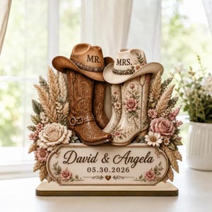 May include: A 2D flat decorative display featuring a pair of cowboy boots and hats labeled MR. and MRS. The brown and cream boots are surrounded by wheat stalks and pink roses. A plaque at the base displays the text David & Angela 05.30.2026.