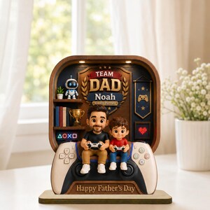 May include: A decorative Father's Day figurine featuring a father and child holding game controllers, seated on a large game controller. The figurine has a wooden frame with the words "Team Dad Noah" and "Happy Father's Day".