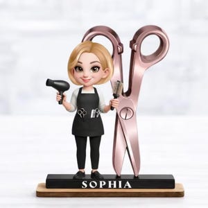 May include: A figurine of a blonde character wearing a black apron over a white shirt and black pants, holding a black hair dryer and a round brush. Beside the character is a large rose gold pair of scissors. The base features the name SOPHIA.
