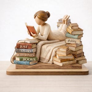 May include: A carved figurine of a person lying on a blanket reading a book, surrounded by stacks of books. The books feature the text "JUST A GIRL WHO LOVES BOOKS" and "#shley". The figure has brown hair in a bun and wears a cream-colored long-sleeved top.