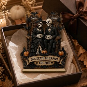 May include: A decorative figurine featuring two skeletons sitting on ornate thrones, holding mugs with heart icons. The piece includes the text Sophia, James, and TILL DEATH DO US PART EST. 2012, accented with bats, pumpkins, and black roses in a gift box.