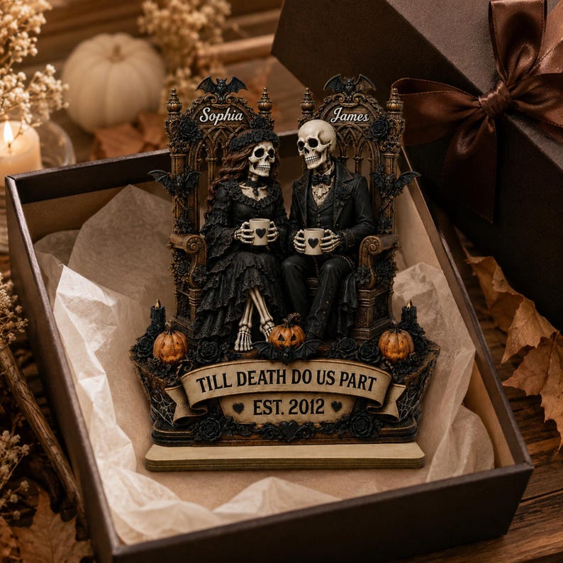 May include: A decorative figurine featuring two skeletons sitting on ornate thrones, holding mugs with heart icons. The piece includes the text Sophia, James, and TILL DEATH DO US PART EST. 2012, accented with bats, pumpkins, and black roses in a gift box.