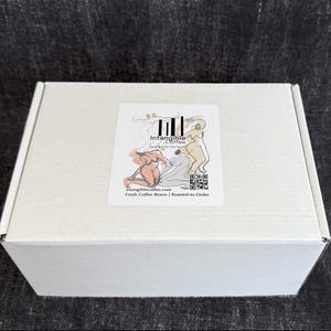 Pu&ograve; includere: Una scatola di cartone bianca con un adesivo con il testo "Intangible Coffee" e illustrazioni di figure. L'adesivo include anche un codice QR e il testo "Fresh Coffee Beans | Roasted-to-Order".