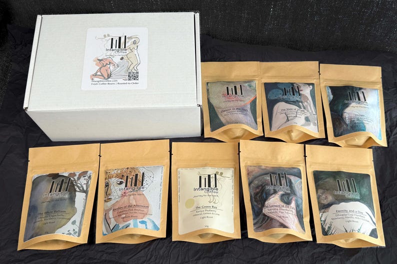 Pu&ograve; includere: Una scatola bianca con un adesivo e diverse bustine di carta kraft con chicchi di caff&egrave;. Le bustine presentano arte astratta colorata e testo. La scatola ha un adesivo con un disegno di due figure e la scritta "Intangible Coffee".