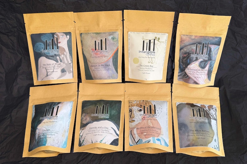 Pu&ograve; includere: Otto sacchetti di caff&egrave; in carta kraft, ciascuno con un'etichetta artistica unica. Le etichette presentano illustrazioni e testo, tra cui "The State of Things" e "The Crown Key". I sacchetti sono disposti su una superficie nera.