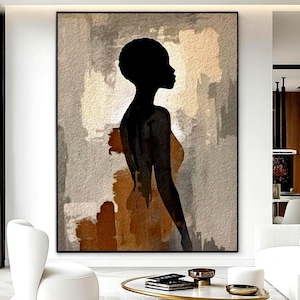 May include: A large framed abstract art print depicting a black silhouette of a person in profile. The background features beige, brown, and grey brushstrokes. The artwork is displayed in a modern living room setting with white furniture.