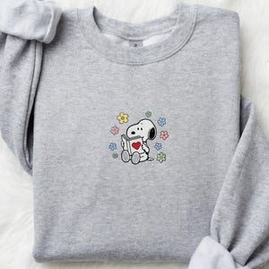 以下が含まれることがあります： 袖をまくったライトグレーのスウェットシャツ。前面には、表紙にハートの付いた本を読むスヌーピーの刺繍が施され、カラフルな花に囲まれています。スウェットシャツは柔らかい素材で作られています。