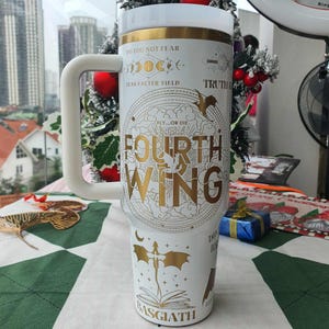 Puede incluir: Taza de viaje blanca con un asa grande, con texto y gráficos dorados y blancos. El texto incluye "FOURTH WING", "BASGIATH" y frases como "DO YOU NOT FEAR". La taza tiene una tapa blanca y una banda dorada cerca de la parte superior.