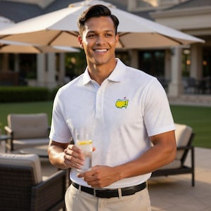 May include: A white polo shirt with a subtle pattern and the Masters logo on the chest. The shirt has short sleeves and a button-down collar. The person is wearing khaki pants and a black belt. The person is holding a glass of water with a lemon slice.