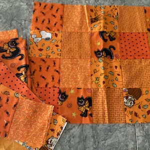 May include: An assortment of orange Halloween-themed fabric squares. The fabric features various patterns, including black cats, Snoopy, candy, and polka dots. The squares are arranged in a patchwork style, with some pieces separate.