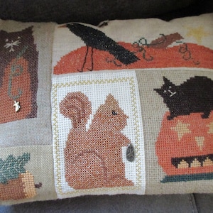 May include: A rectangular throw pillow featuring a cross-stitch design on a tan fabric background. The pattern includes a black cat, a crow on an orange hill, a brown squirrel, a pumpkin with a black cat, and an acorn, accented with small metal charms.