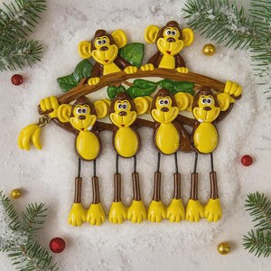 May include: A whimsical monkey ornament featuring six smiling monkeys in shades of brown and yellow. The monkeys are hanging from a brown branch with green leaves, with a bunch of yellow bananas. The ornament is decorated with festive holiday accents.