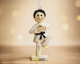 May include: A karate-themed ornament featuring a figure in a white uniform with a black belt, poised in a martial arts stance. The ornament has a smiling face and a small base with the text "1 NAME" and "2 YEAR". A white ribbon is attached for hanging.