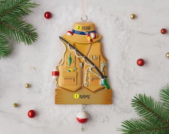 Könnte beinhalten: Ein Weihnachtsornament in Form einer Fischerweste, eines Hutes und einer Angelrute. Die beige Weste hat Taschen und eine Angelrute. Der Hut trägt den Text "2 YEAR". Das Ornament hat eine Pose mit dem Text "1 NAME".